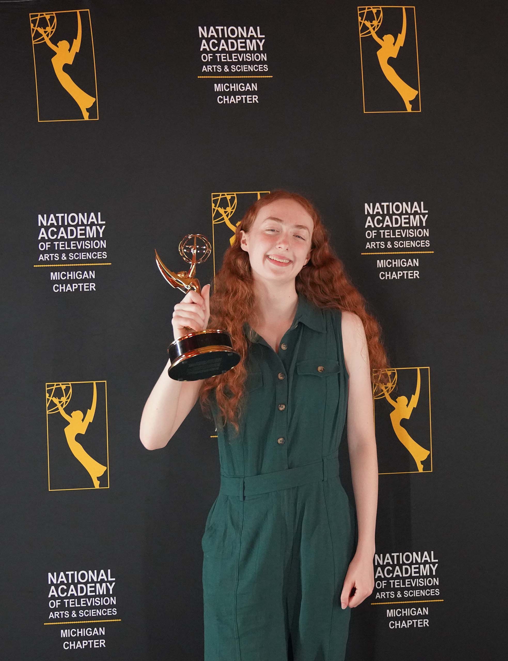 Attired in a green jumpsuit, Renata Schmult smiles and holds up her Emmy in front of a banner with logos for the National Academy of Television Arts & Sciences Michigan Chapter.