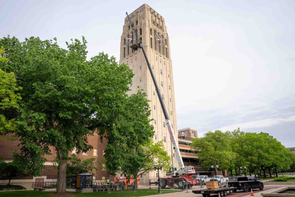A crane reaches up to the top levels of Burton Memorial Tower, and to an exterior platform fixed to the bell chamber windows. On the ground, fencing and facilities trucks surround the tower.