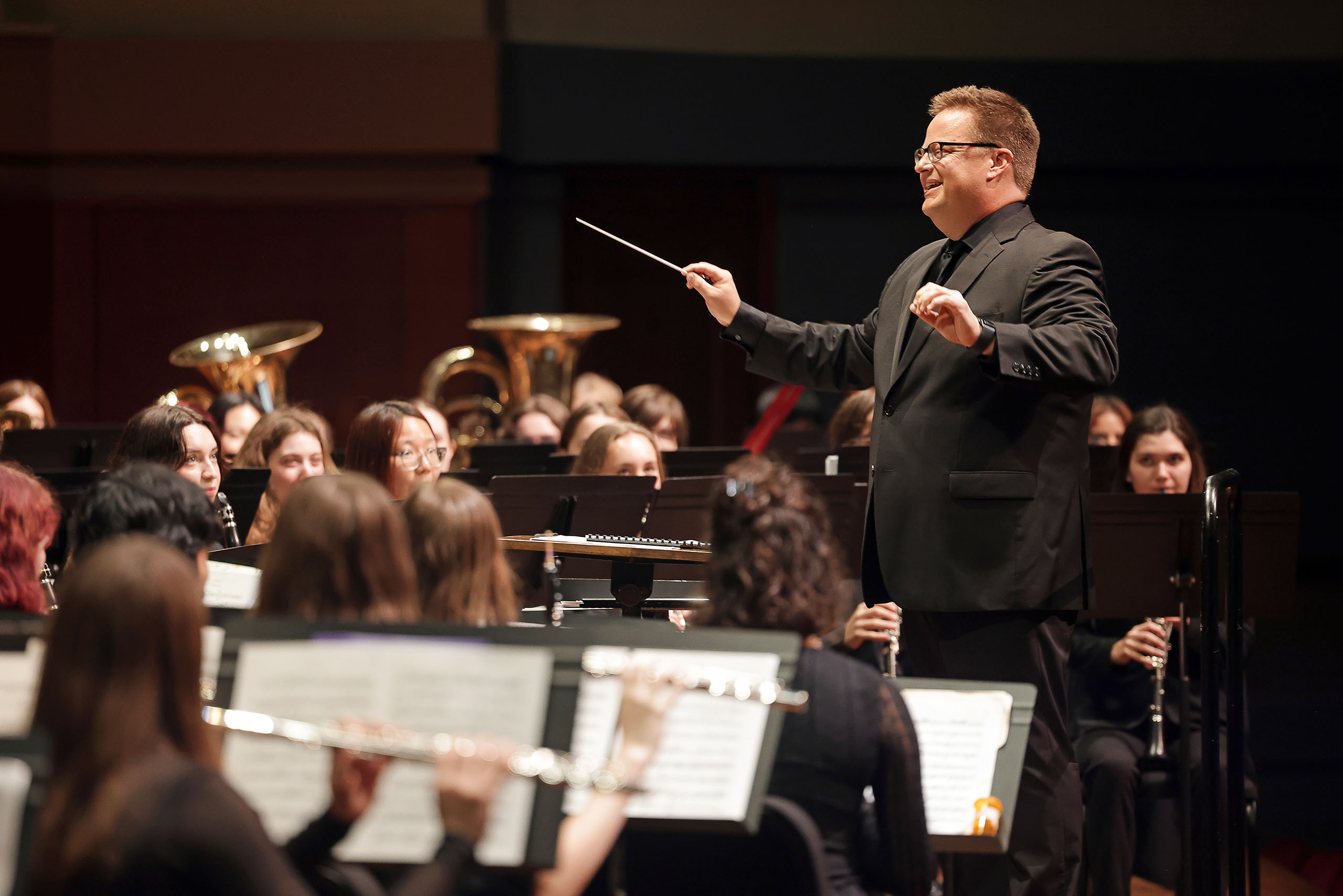 Richard Frey conducts a band in a performance, with his baton tipped up, wearing formal black concert attire.