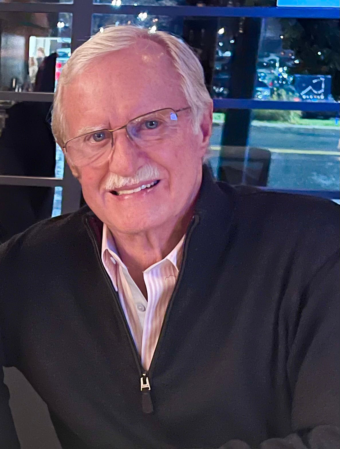 Casual headshot of Richard Grunow seated, with a city street outside the window behind him.