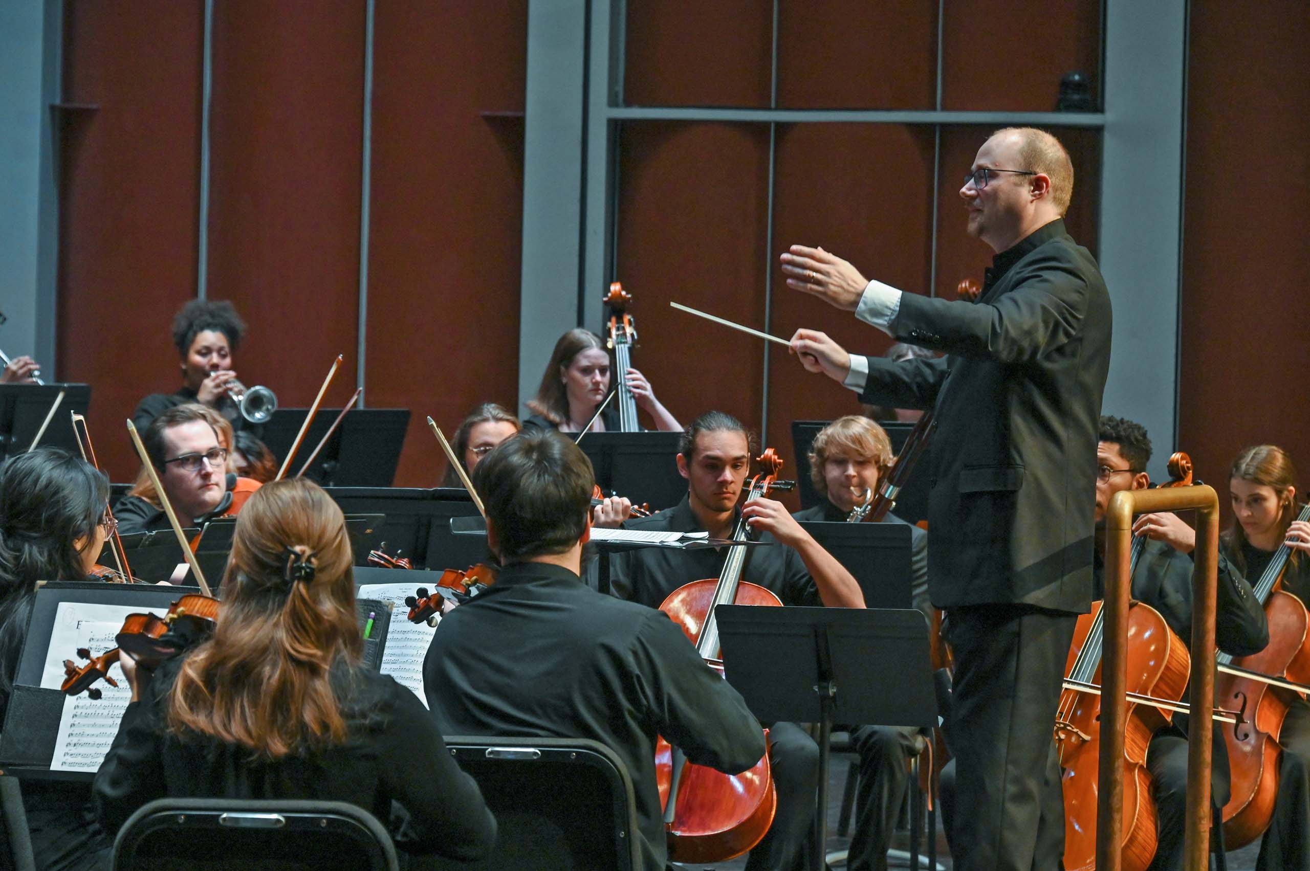 Rotem Weinberg stands at a podium and conducts an orchestra performing in concert with arms upheld forward.