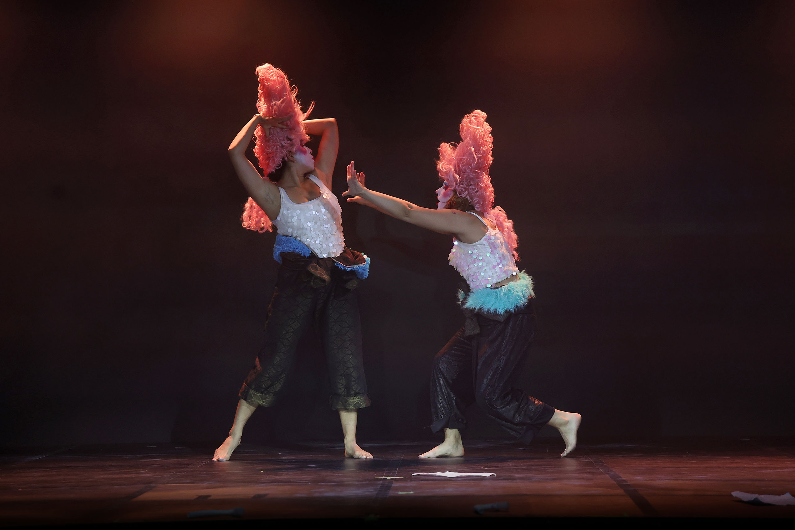 Two dancers perform on a dark stage costumed in black pants, white sequined tops, tall pink wigs with curls, and white makeup. One dancer holds their arms pushed out toward the other dancer, whose arms are bent back behind their head.