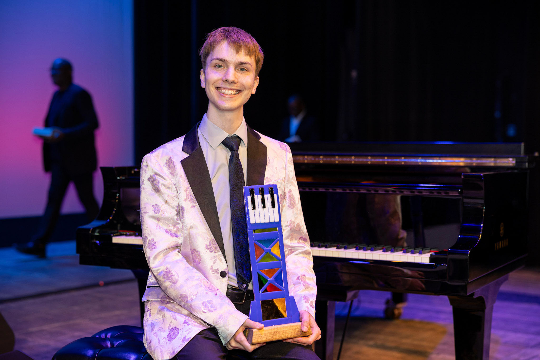 Seated on a stage in front of a piano, and wearing a suit coat, Rowan Tucker-Meyer smiles and holds a colorful trophy featuring piano keys.