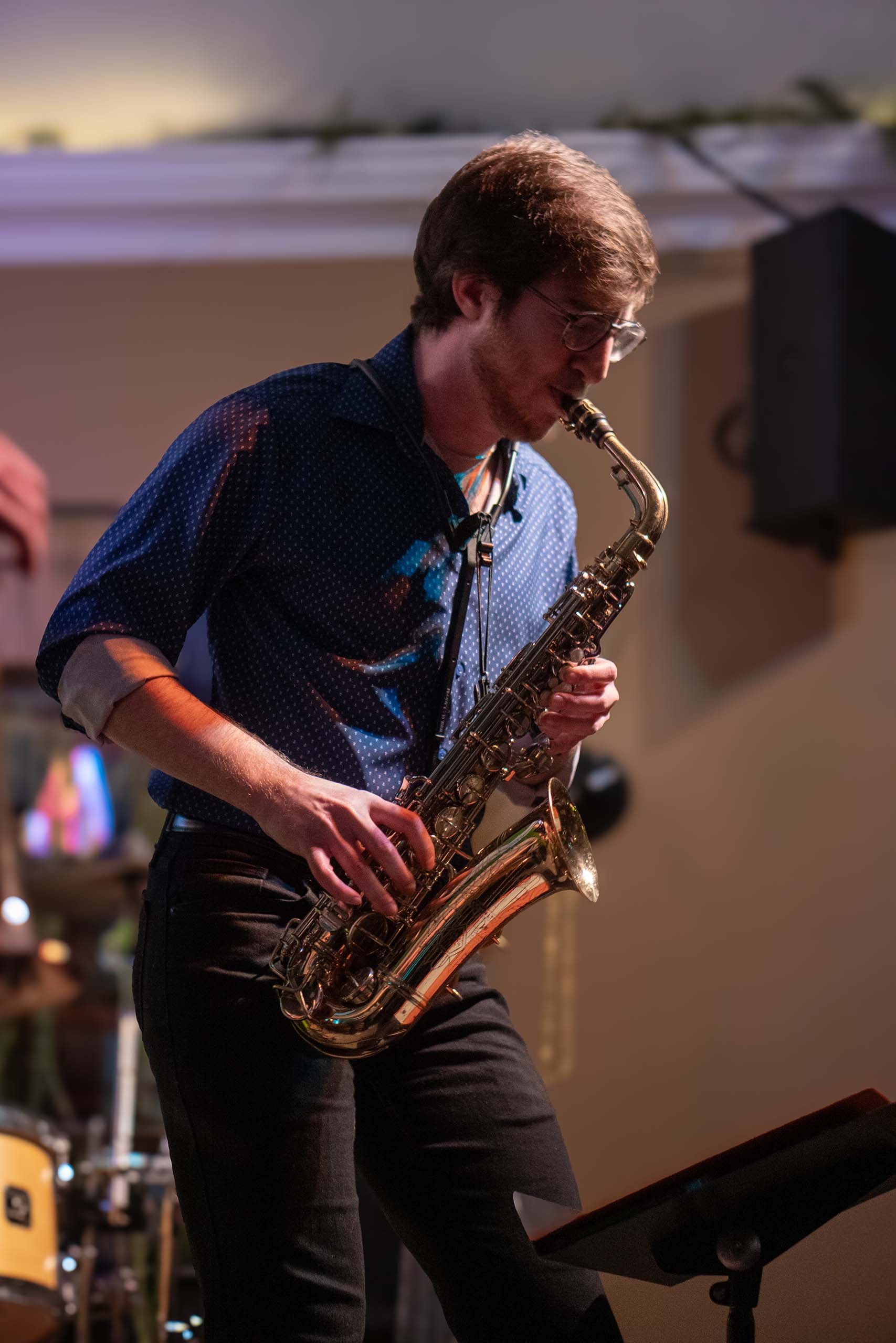 Ryan Schildcrout stands performing saxophone on a small stage, attired casually.