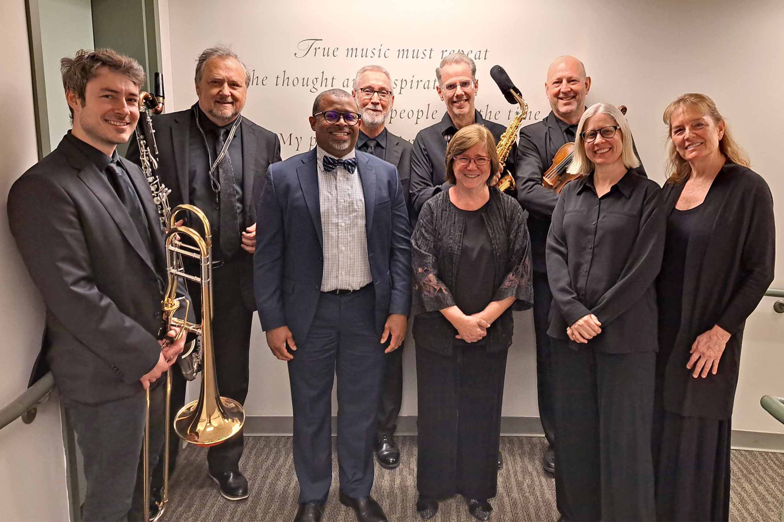 Nine people pose standing together in two rows, wearing formal attire. Four of them hold instruments.