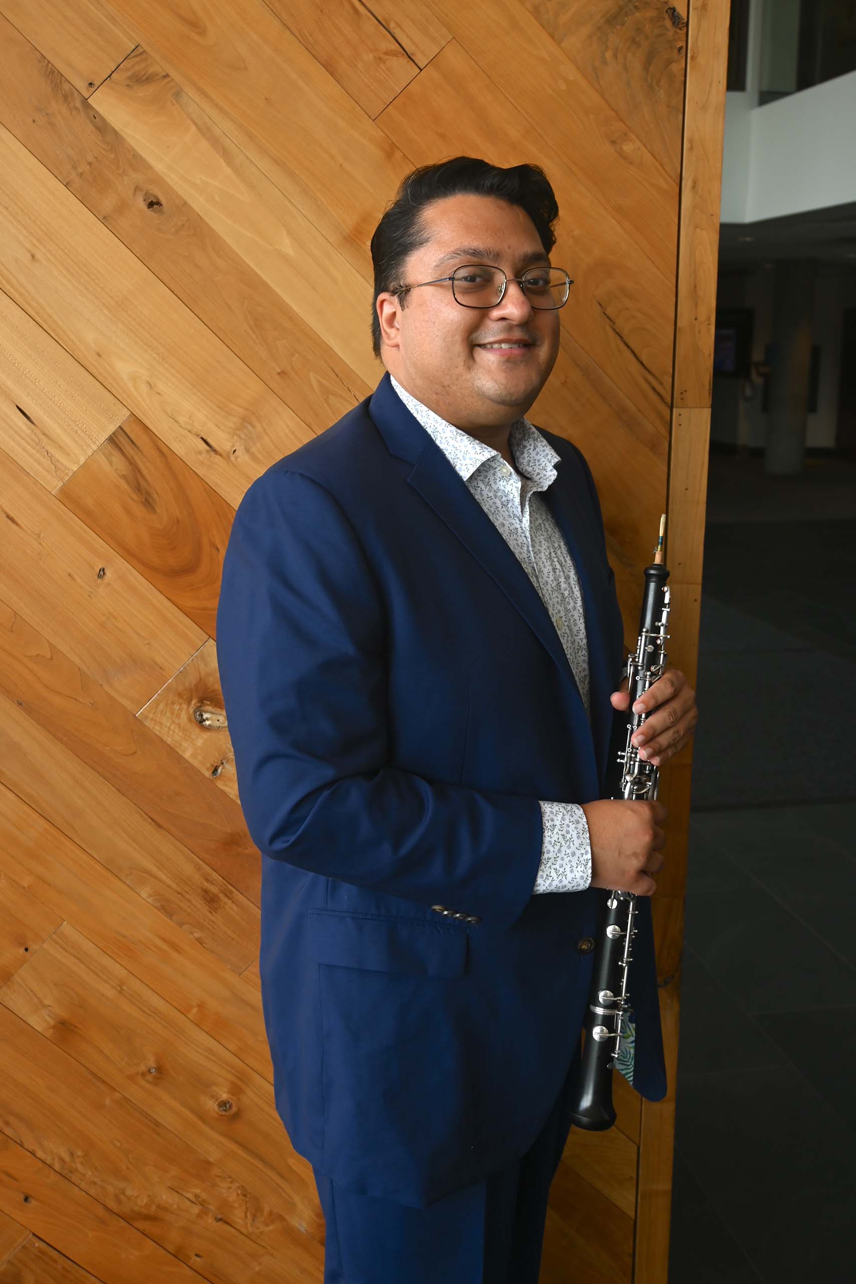 Portrait of Sagar Anupindi standing and holding an oboe, attired in a navy blue suit, in front of a wood-paneled wall.