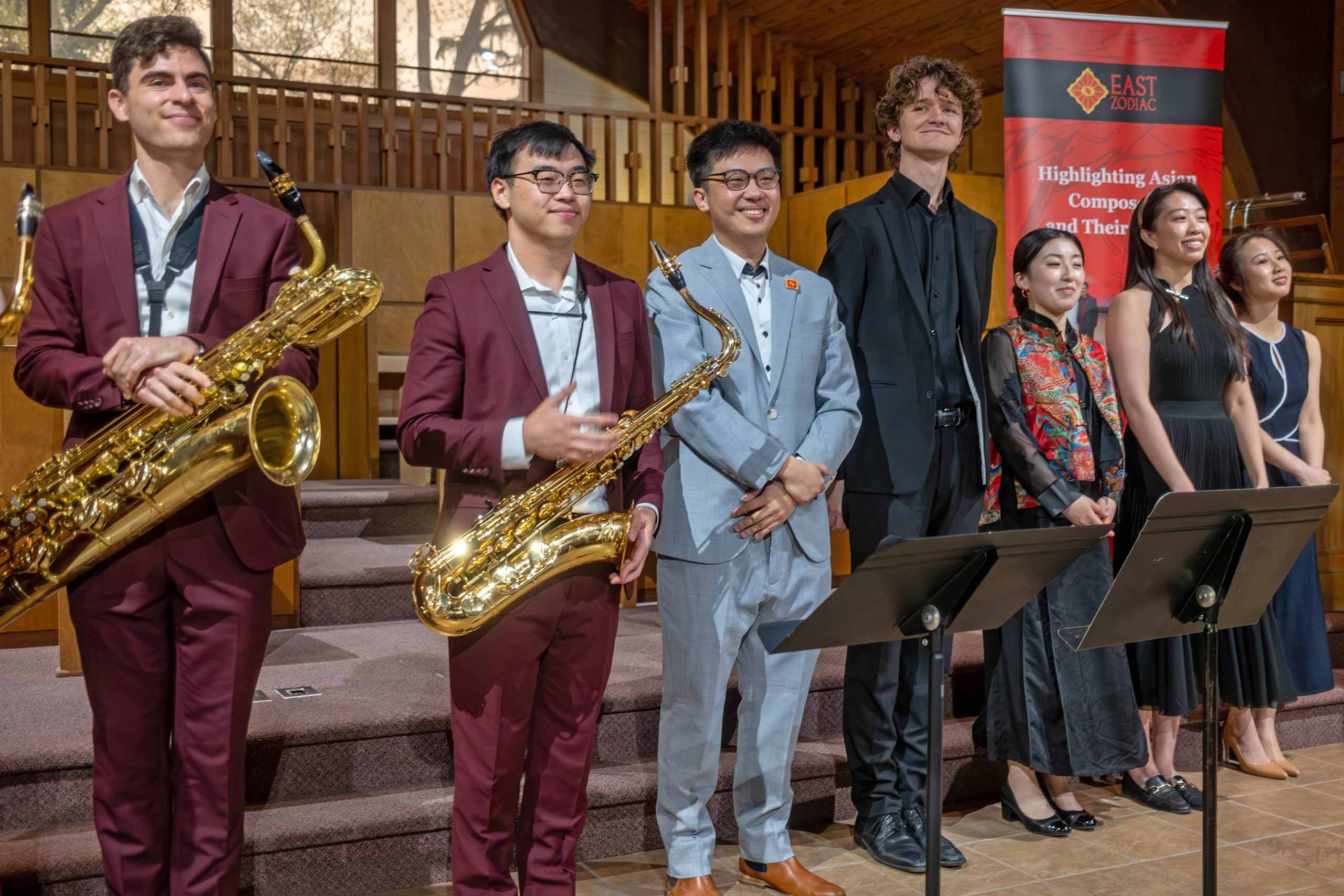 Seven people stand side-by-side smiling with music stands at the front of a church venue, wearing concert attire. Two of them are dressed alike in burgundy suits and hold saxophones.