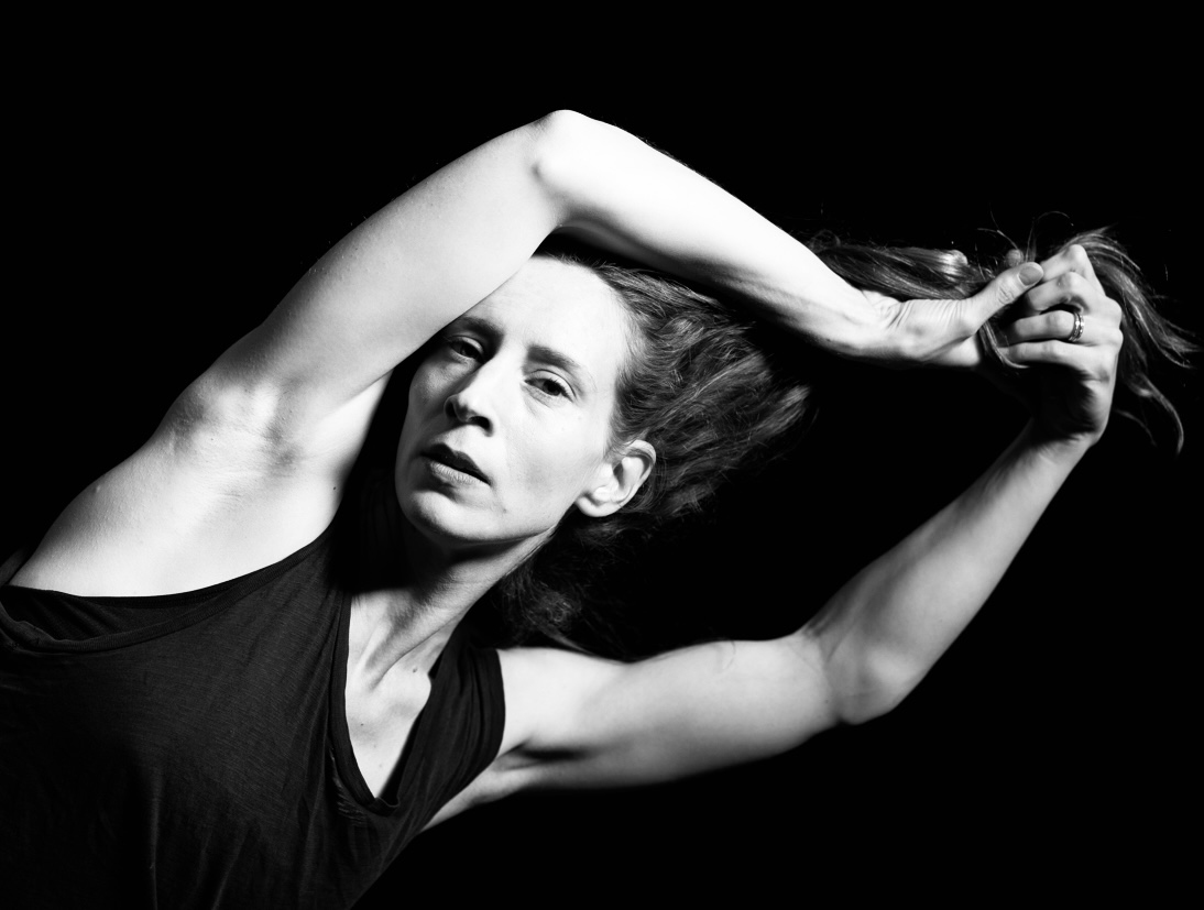 Greyscale portait of Shannon Gillen. Attired in a simple black tank, she leans sideways with arms raised, holding her hair out at the ends.