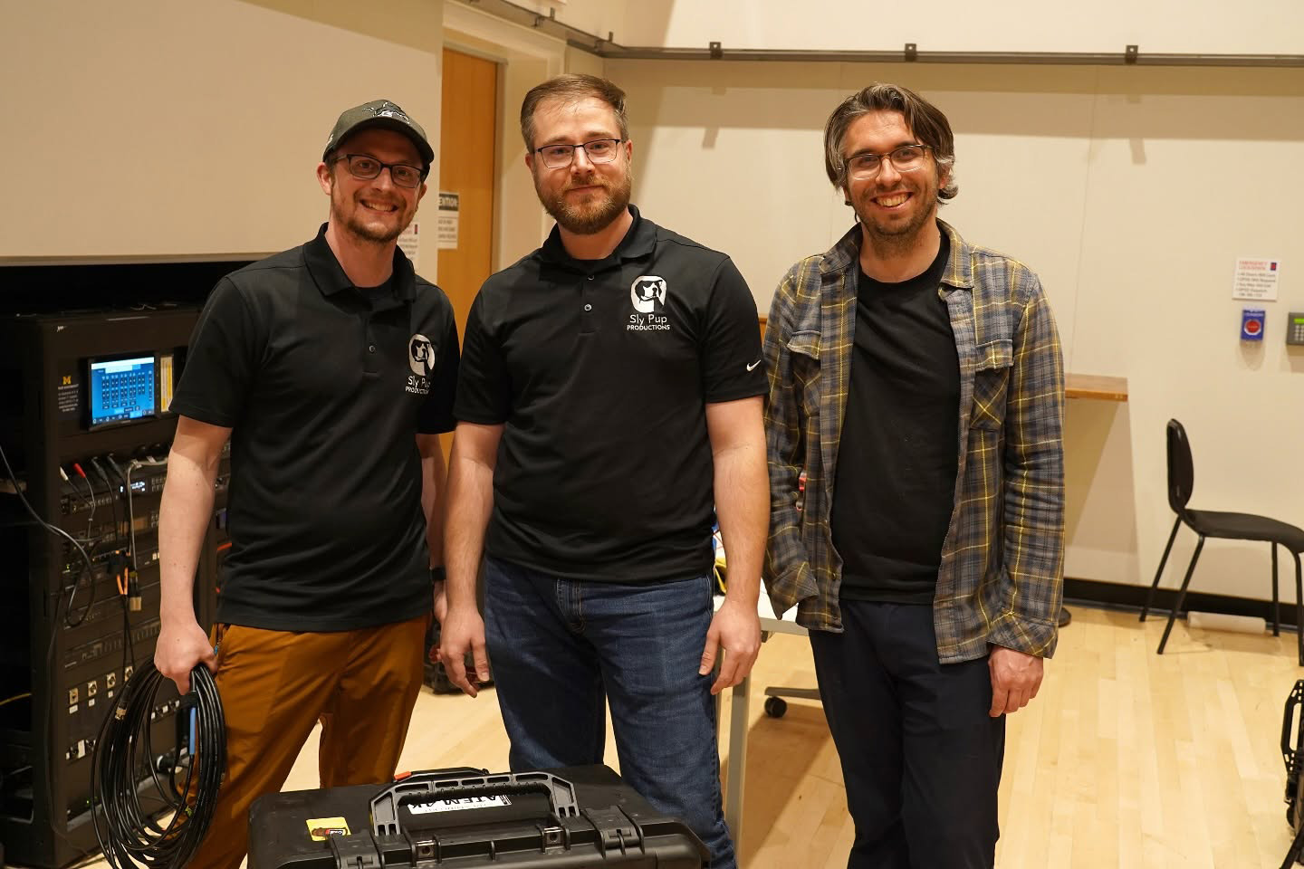 Three people pose together standing in front of a production console in a studio space. One holds a bundle of audio cables and two are wearing black t-shirts with the SlyPup logo.