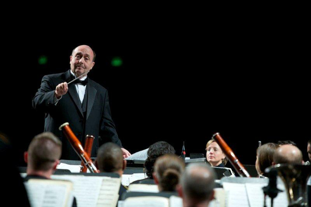 Stephen Pratt conducts in a dark hall, attired in a concert tuxedo. In the foreground, musician heads and bassoons peer above sheet music on stands.