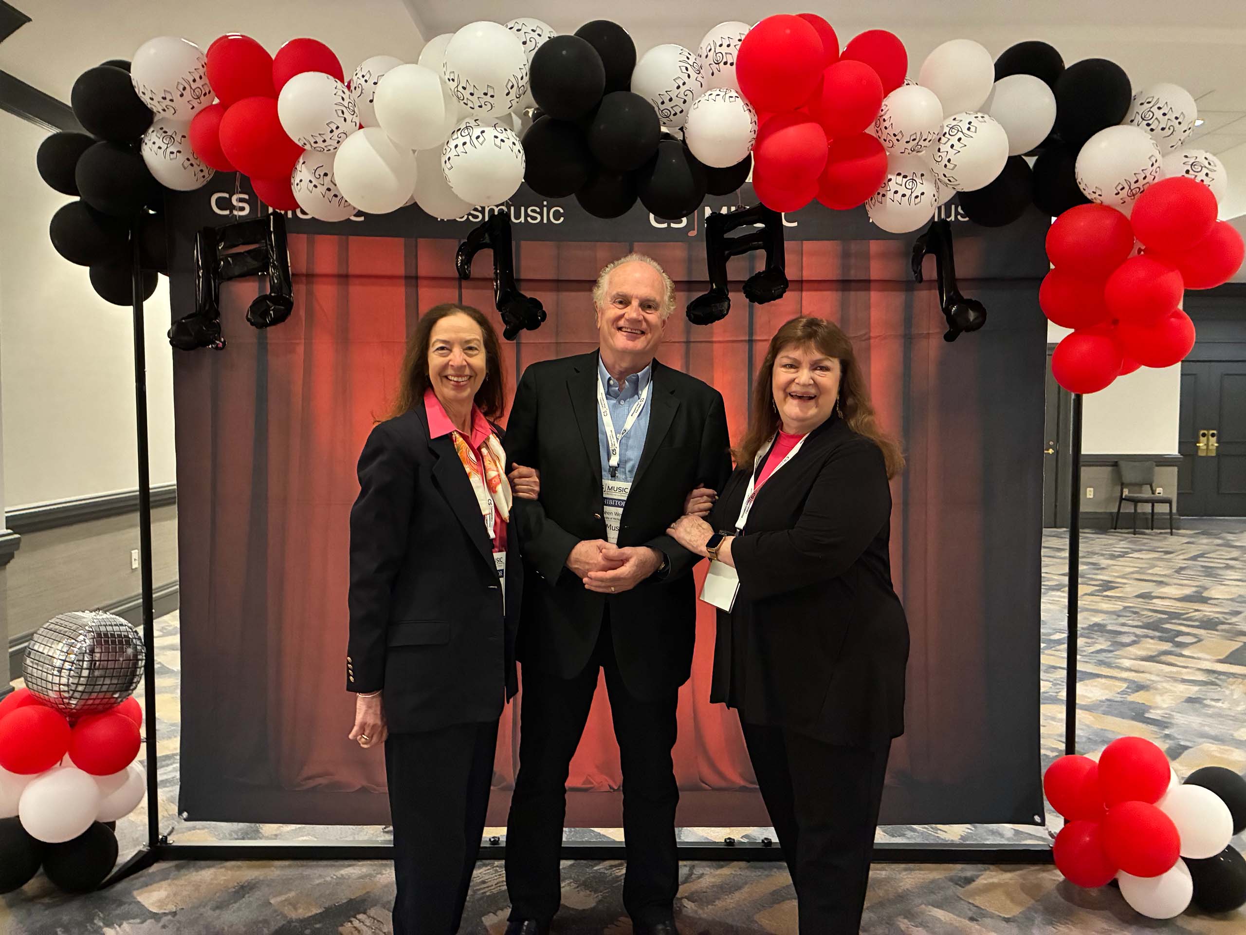 Three people pose standing together, all wearing suits and conference nametags on lanyards. They are in front of a photo backdrop featuring a red curtain graphic, music notes, and balloon clusters in red, white, and black.