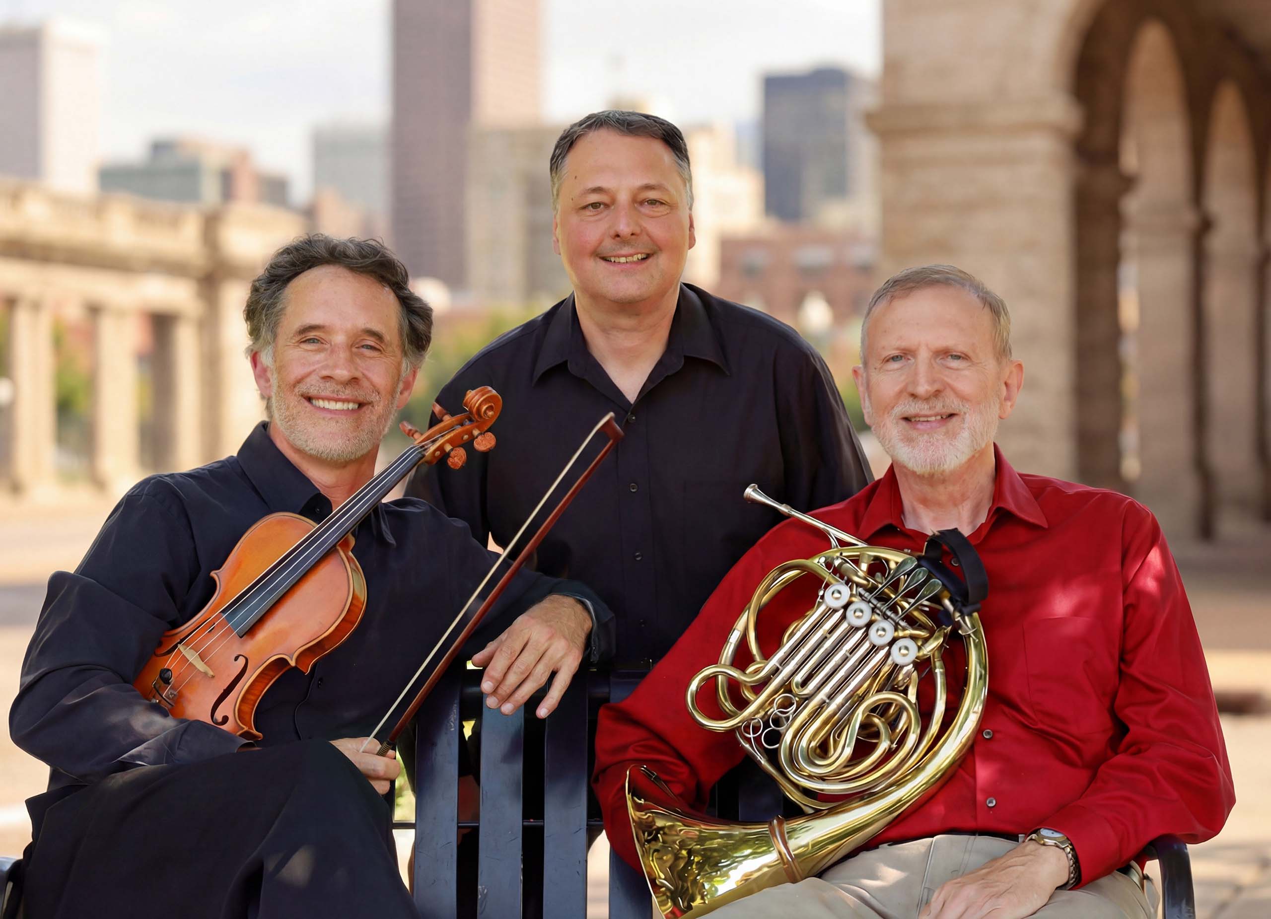 Group portrait of three people with a cityscape in the background. One holds a violin and one holds a French horn.