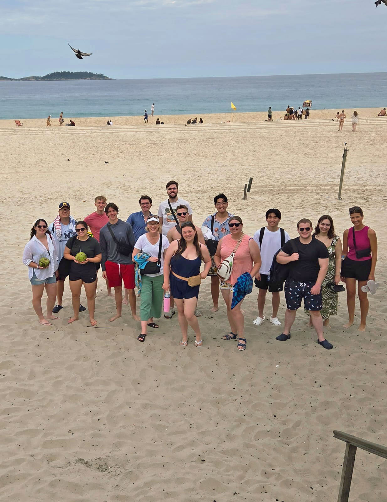 Sixteen people pose together standing on a wide beach, in casual beach attire, looking up at the camera. The waterfront behind them includes other beachgoers, a bird in the sky, and land with trees rising in the distance of the horizon.