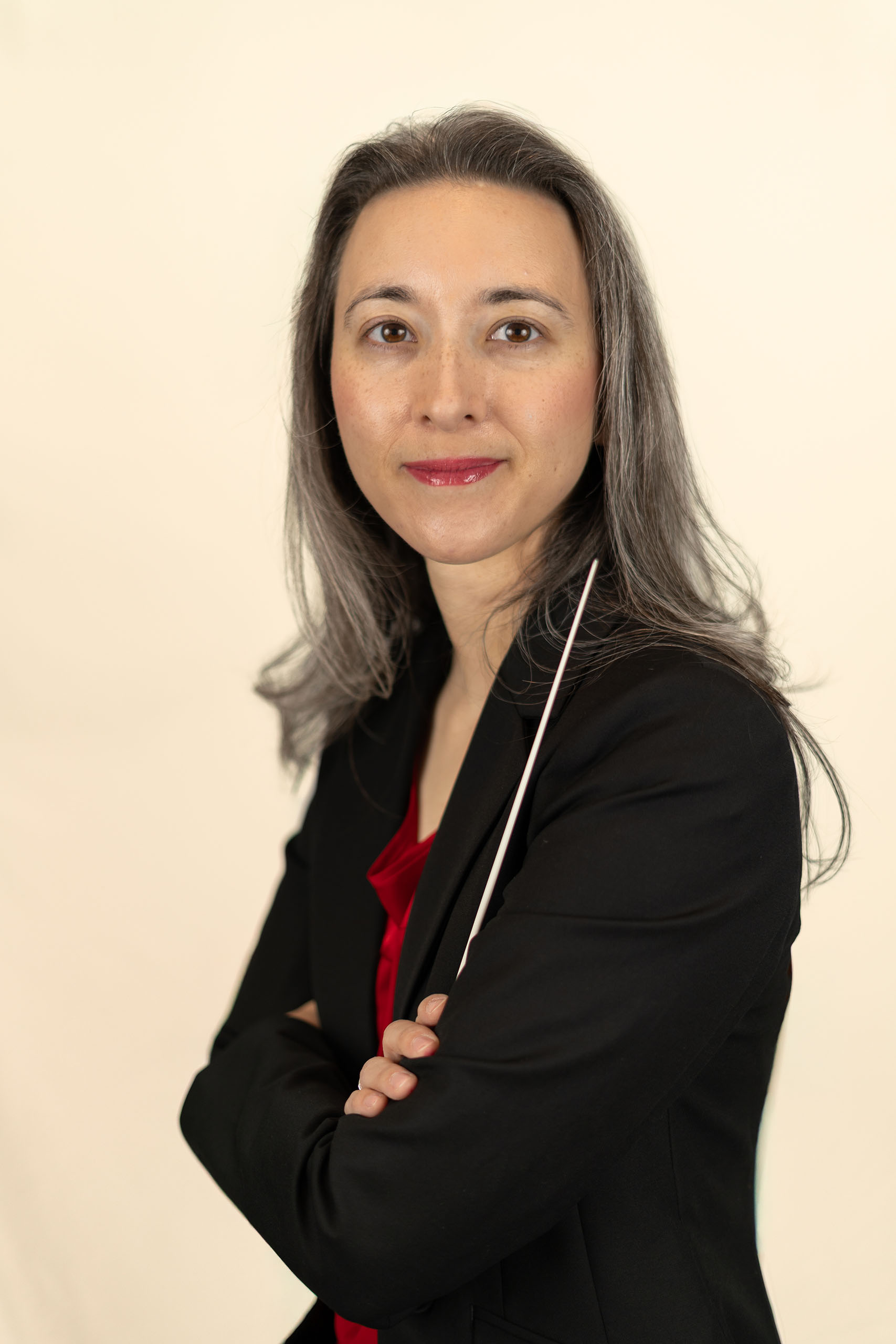Portrait of Taryn Wood wearing a black suit coat, facing to the side with arms folded and holding a conductor's baton.