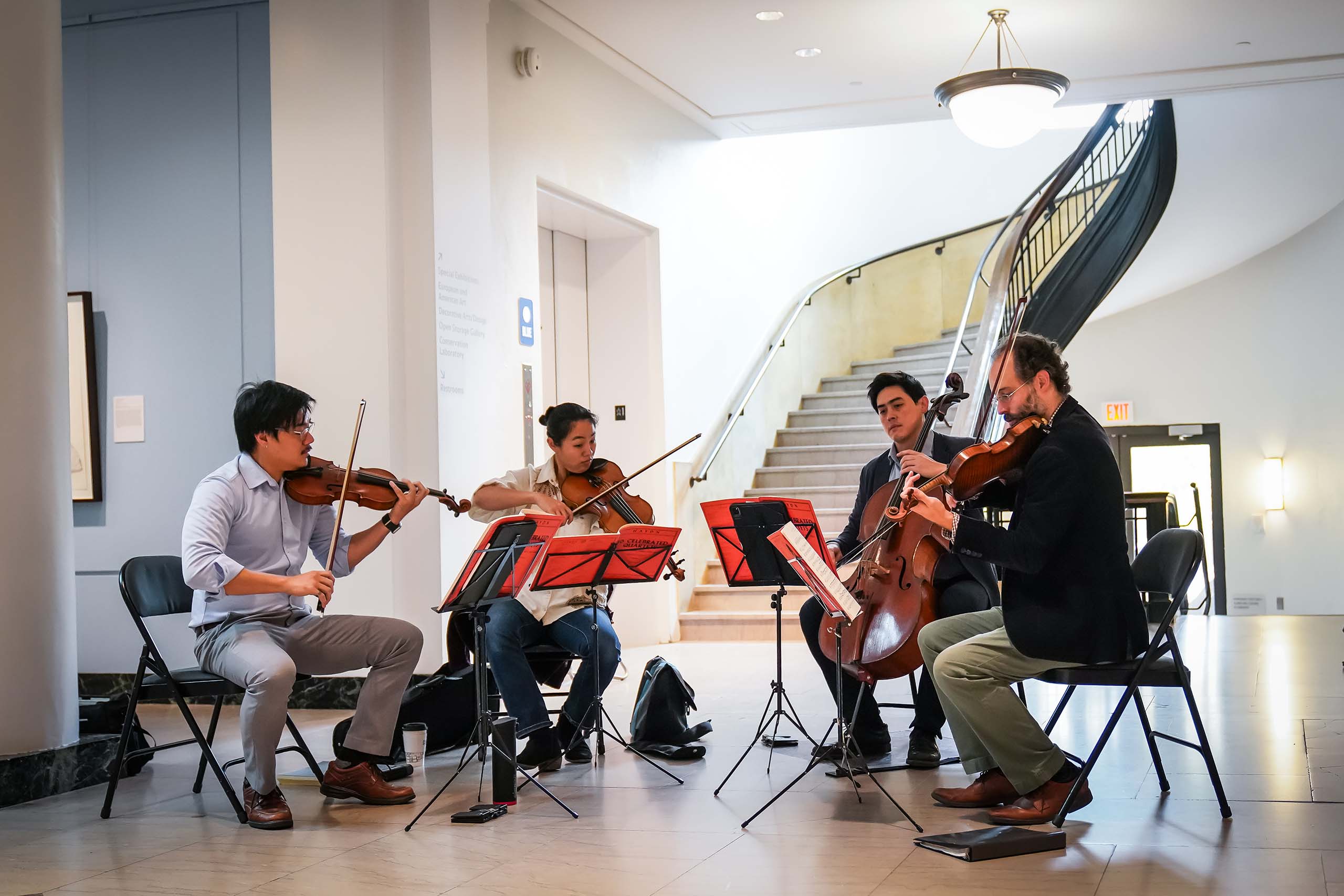 A seated string quartet performs together using portable music stands and folding chairs. Around them we see the light marble floors of the U-M art museum, with the elevator and a curved stairwell in the background.