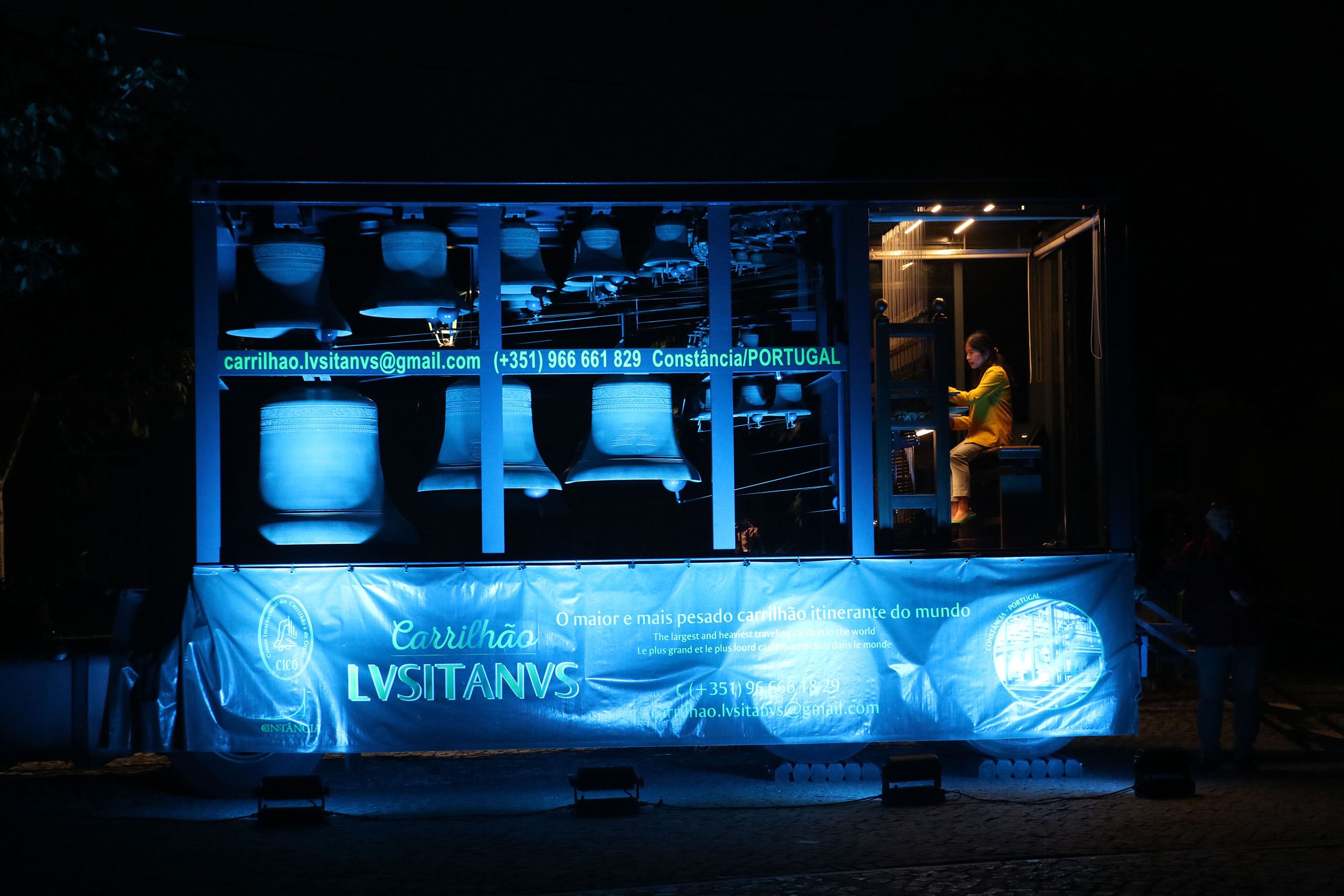 Tiffany Ng performs on a mobile carillon, with two tiers of bells the size of a large trailer. A banner with foreign language covers the trailer wheels. The carillon is lit with blue light, while Tiffany is under white lights, in an otherwise pitch black area.