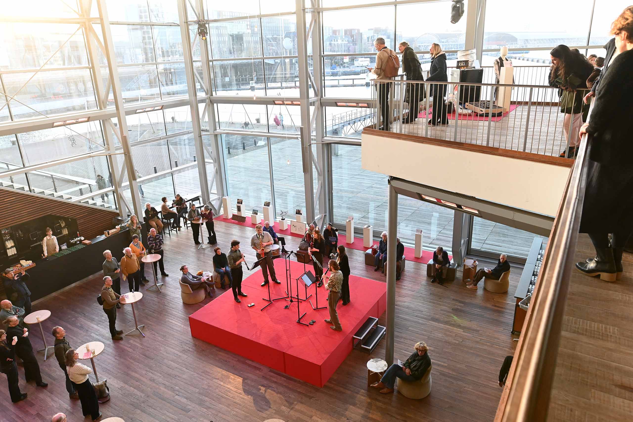 A wind quintet performs standing on a raised red platform stage, in the middle of a two-story lobby space with modern glass walls, steel structure and interior balcony. Over two dozen viewers of the performance are visible facing the stage, both seated and standing at bistro tables.