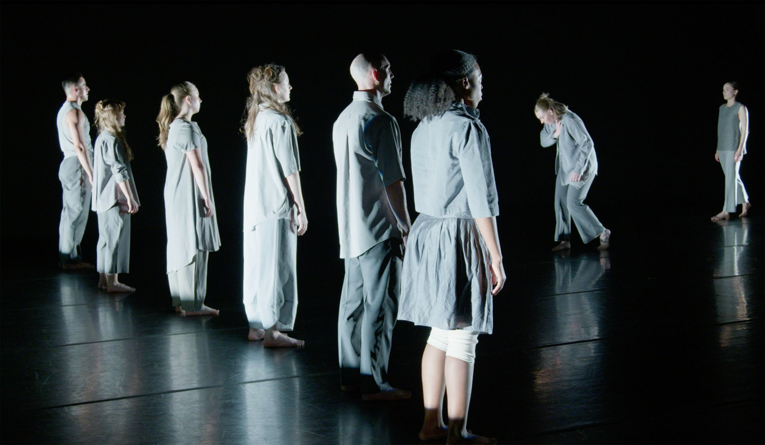 On a dark stage, six dancers stand side-by-side while two others face them in movement. All are costumed in loose-fitting, light grey casual attire.