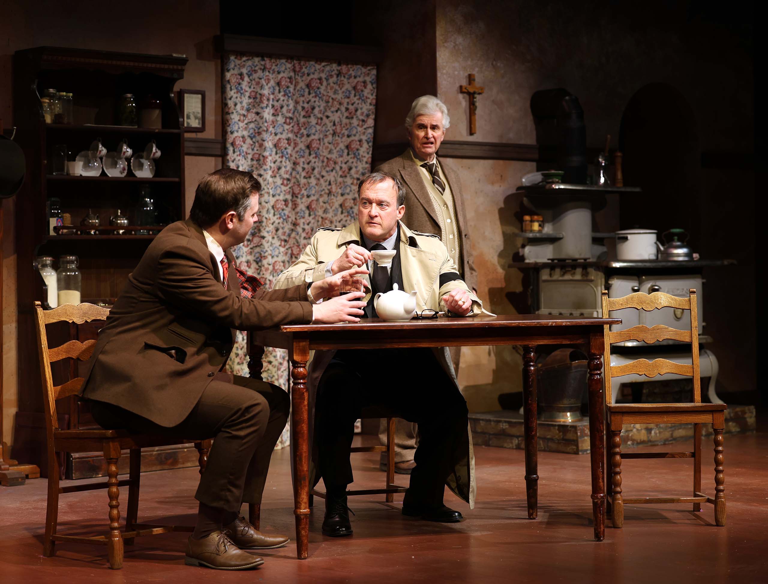 Three actors perform a scene on stage around a kitchen table set in the early 20th century. They are costumed in brown and neutral toned suits and jackets.