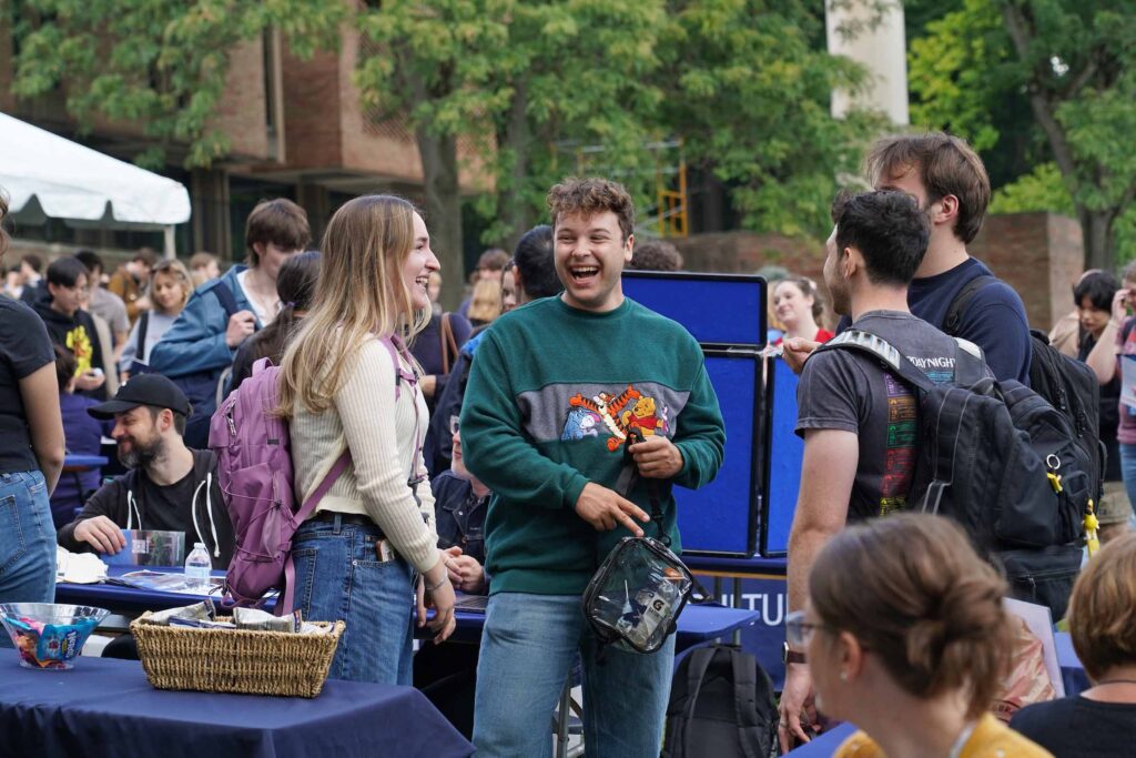 A group of four students stand together talking, with three wearing backpacks. They stand amidst a crowd and display tables at the Welcome Back Carnival.