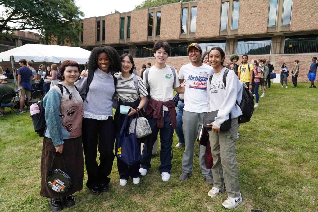 Six students pose standing together and smiling, all with backpacks, outside the Moore Building at the Welcome Back Carnival.
