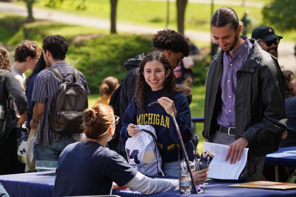 Two students stand interacting with someone seated at a table outdoors at the Welcome Back Carnival. One wears a Michigan shirt and holds an SMTD cinch bag.