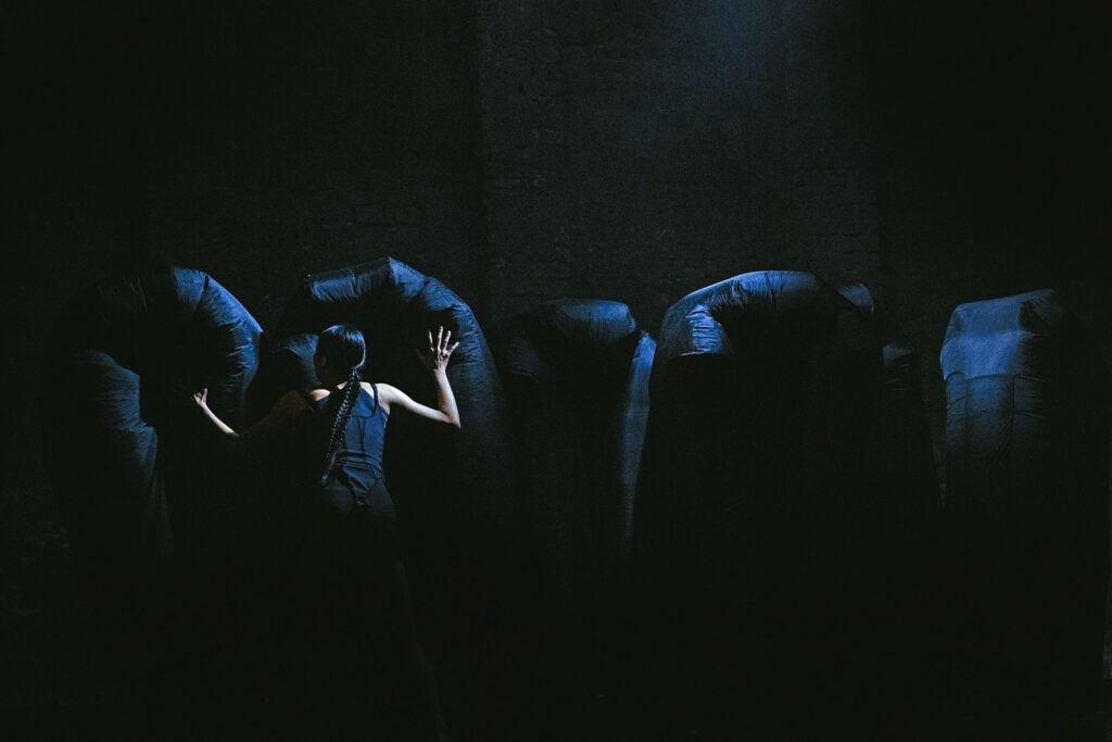 In a dark performance space, a dancer attired in black faces a series of black shapes filled with air.