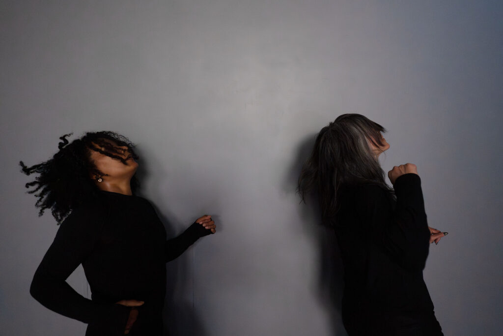 Two dancers attired in black long-sleeved shirts against a gray wall.