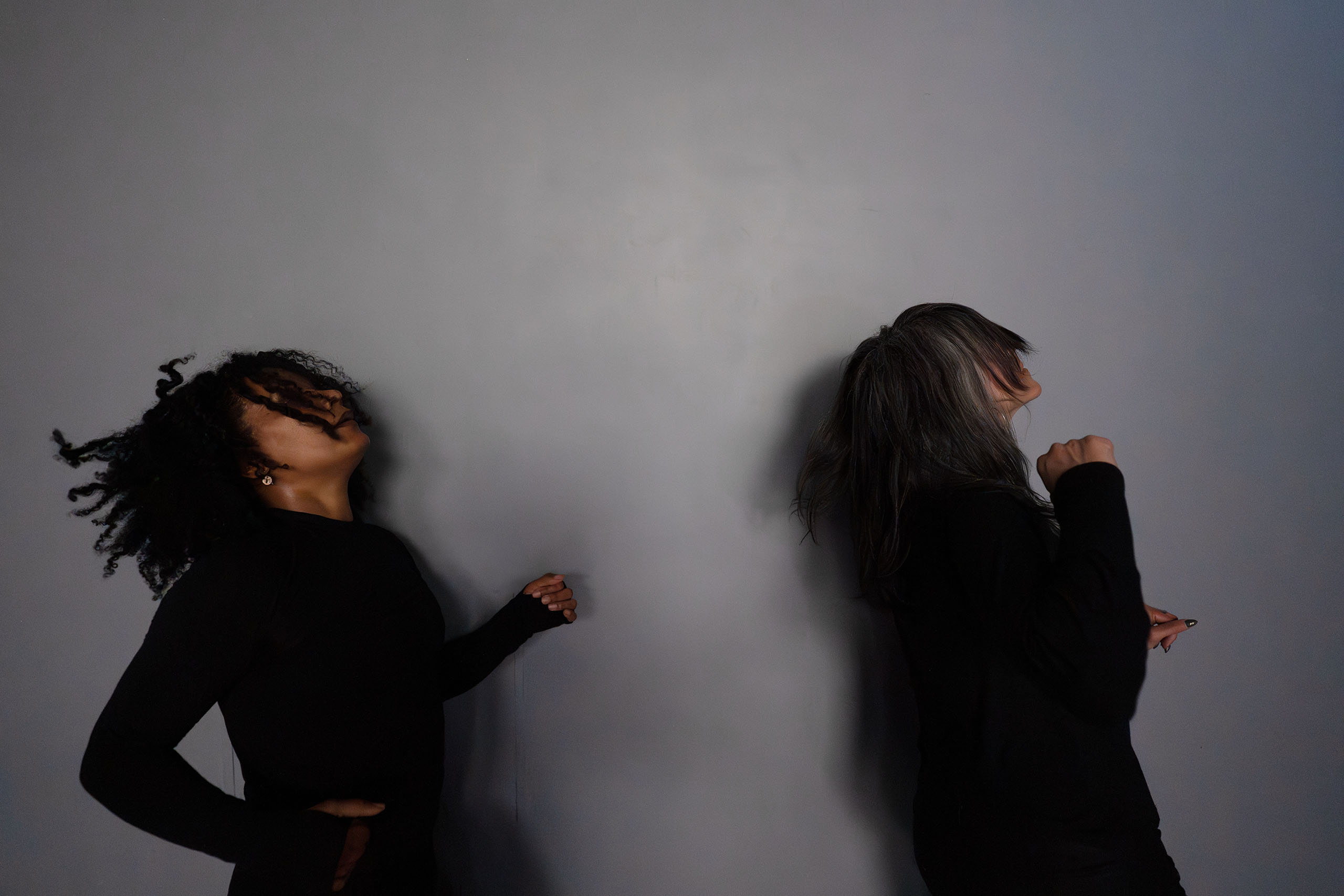 Two dancers attired in black long-sleeved shirts against a gray wall.