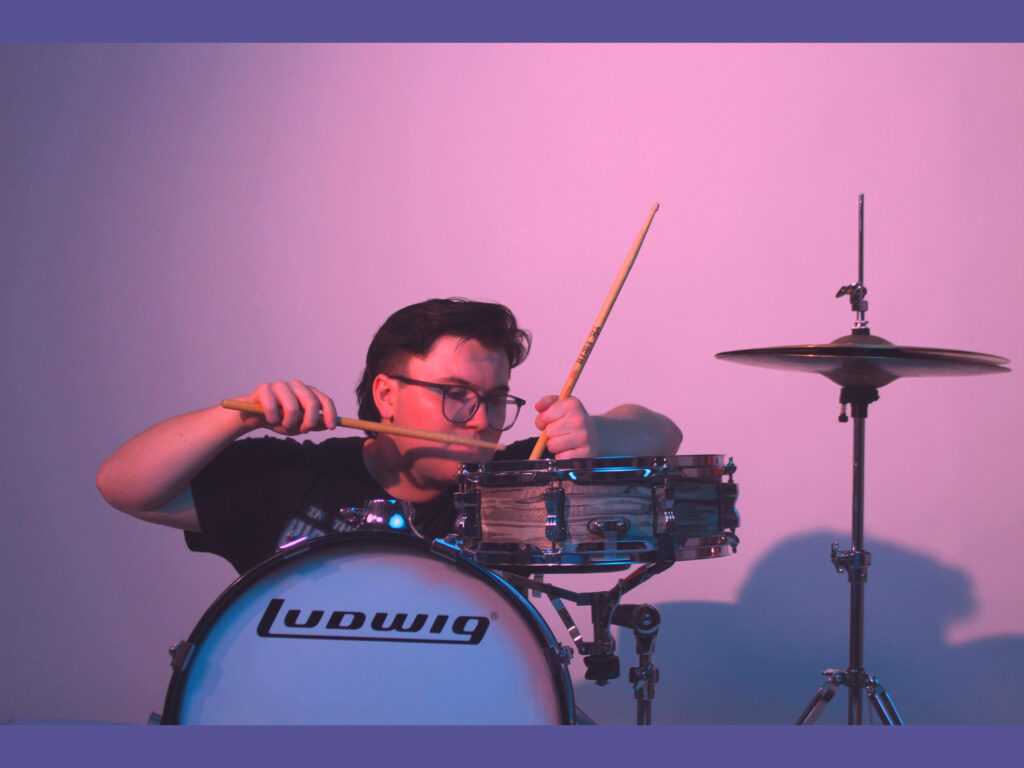 Portrait of Elijah Hall playing a snare with drumsticks, with a purple background.