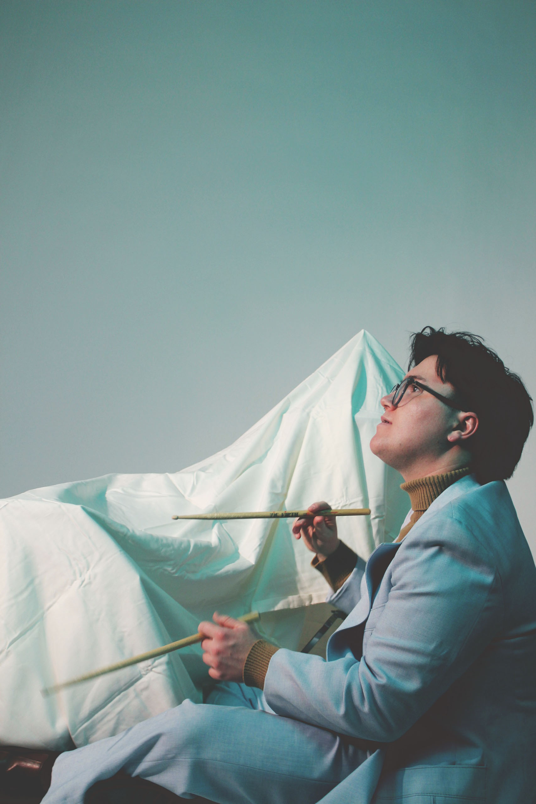 Candid portrait of Elijah Hall holding drumsticks in both hands; attired in a light blue suit. His drum set is covered in a sheet behind him.