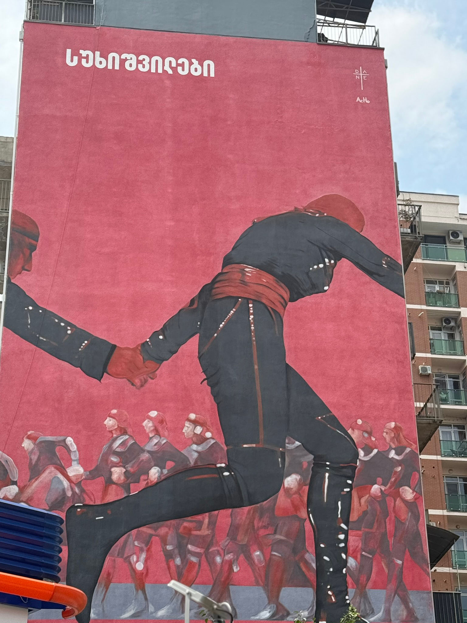 A mural painted on a windowless building surface about 8 stories tall. In the mural, people wearing black and red full-body uniforms hold hands, with a red background and white lettering at the top in a foreign language.