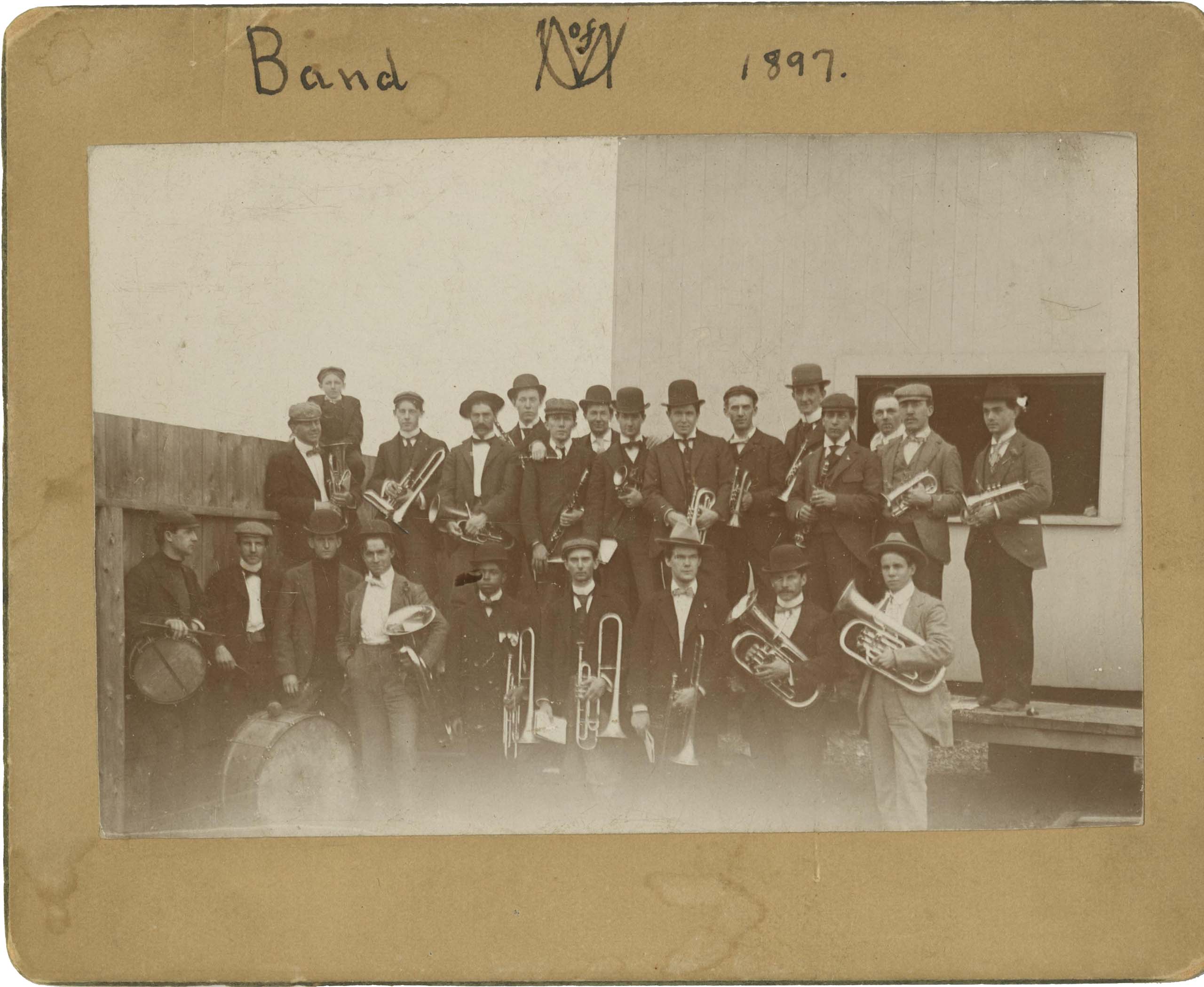 Old black and white photo of 23 band members in two rows, holding instruments and wearing suits and top hats; mounted to a board with "U of M Band 1897" written by hand.
