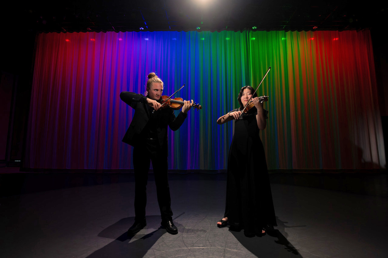 Two violinists stand performing in black attire, with rainbow lighting on a curtain in the background.