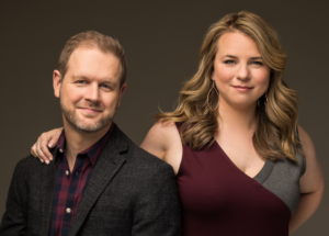 The photo shows two people posing together against a plain background. The man on the left has short light brown hair and a trimmed beard, wearing a dark textured blazer over a red and navy plaid shirt. The woman on the right has long, wavy blonde hair, is wearing a sleeveless dress with maroon and gray sections and large hoop earrings, and has her arm resting on the man's shoulder. Both are smiling softly at the camera.