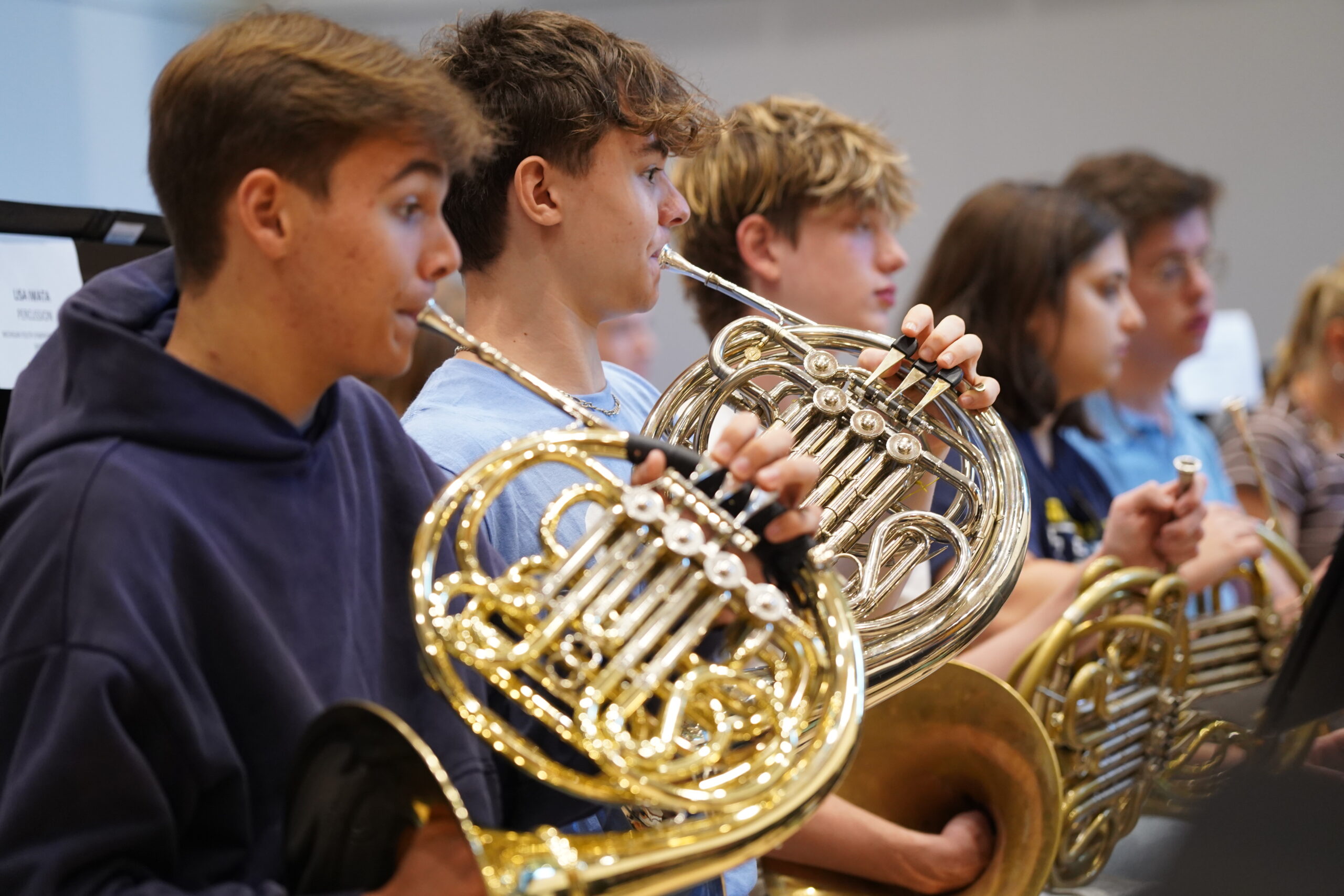 A row of 4-5 students playing French horns seated in a rehearsal.