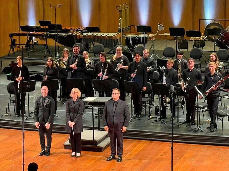 On a performance stage, three leaders stand in front of a conductor's podium with 14 wind players behind them; all are standing for a bow attired in concert black.