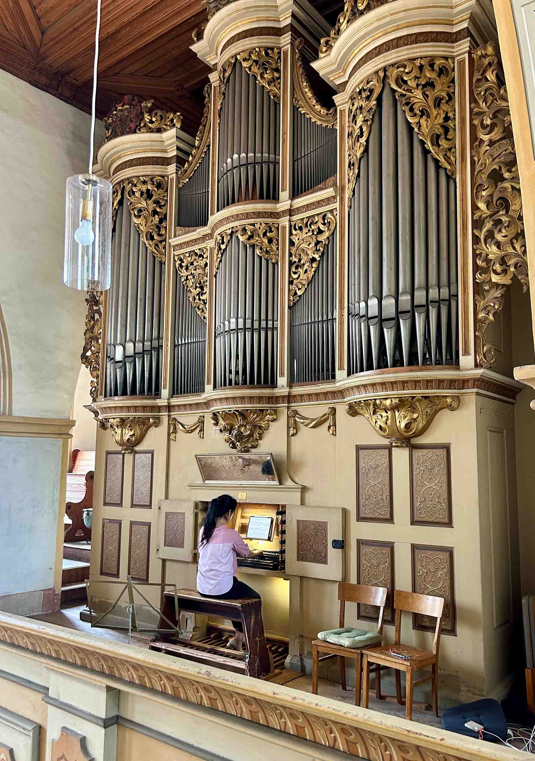 Chamni Kim plays a two-story historic organ with a small console, with painted decorations and ornate gold trim around the pipes.