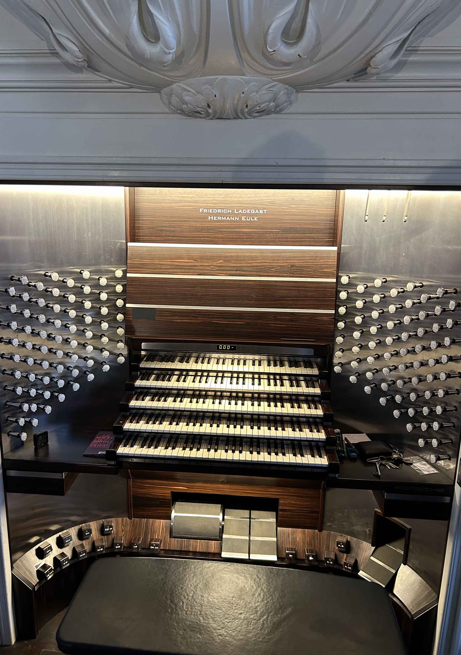 A modern organ console with curved interior walls made of steel and wood panels, with inset lighting over the keyboards, pistons, and foot pedal areas.