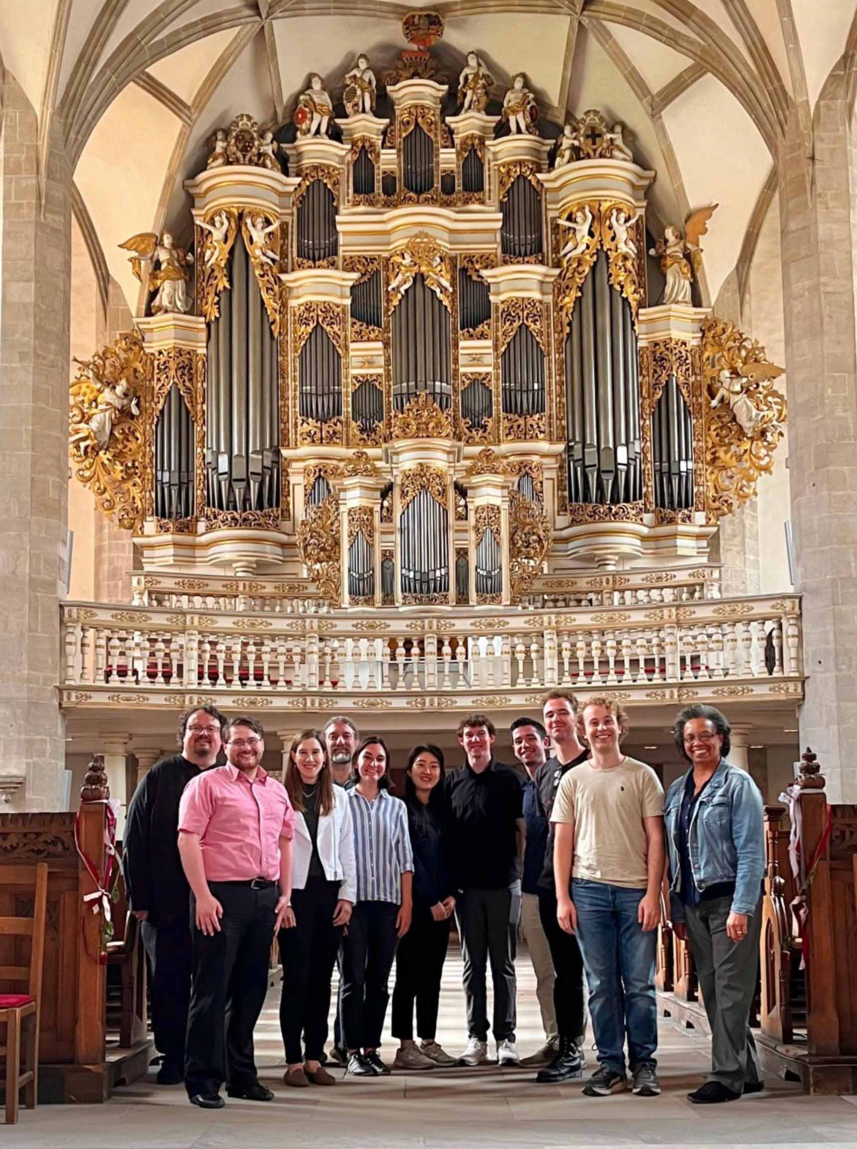 Eleven people pose standing together in a large historic church, with ornate multi-story organ pipes behind them.