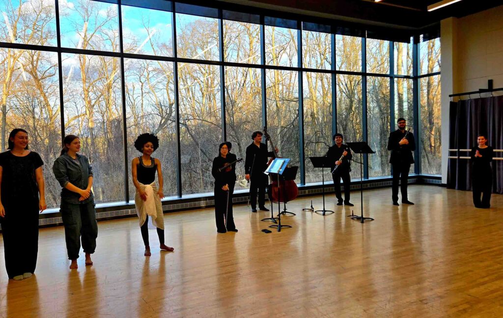 Dancers and musicians stand for a bow in a studio with large windows overlooking woods.