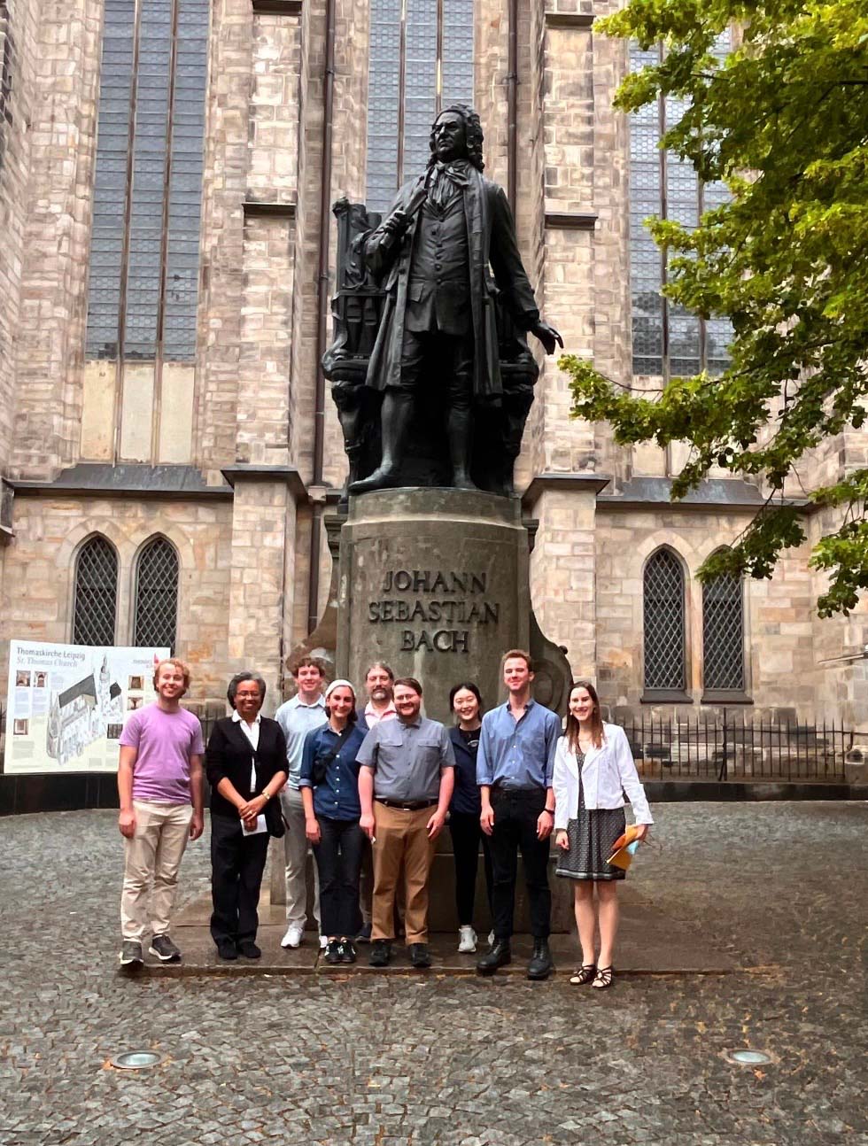 A group of nine people poses standing outdoors, next to a bronze statue on a pedestal, labeled Johann Sebastian Bach. An informational display and a historic church are in the background, and the ground is paved with stone.