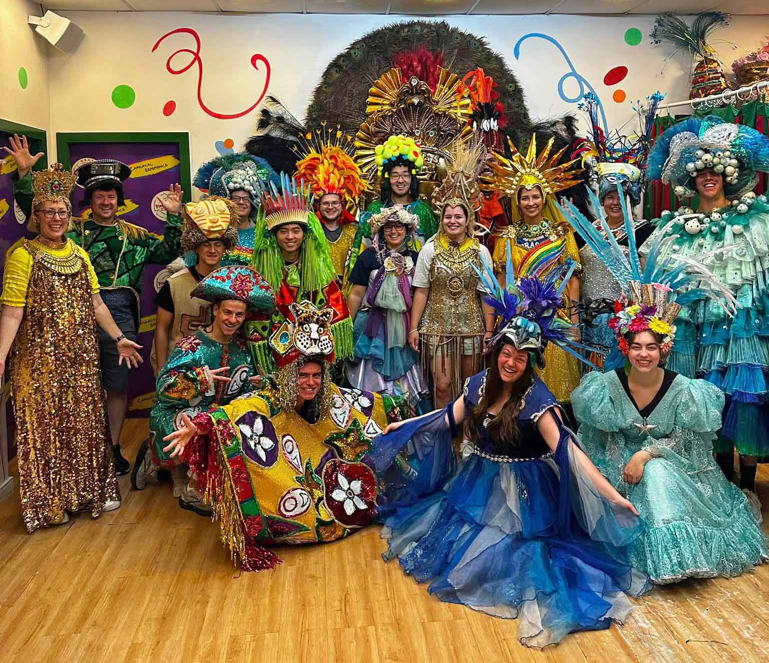 About 16 people pose as a group, all are attired in Brazilian costume featuring bright colors, long draping fabrics, and elaborate headwear. Many hold out their arms and hands.