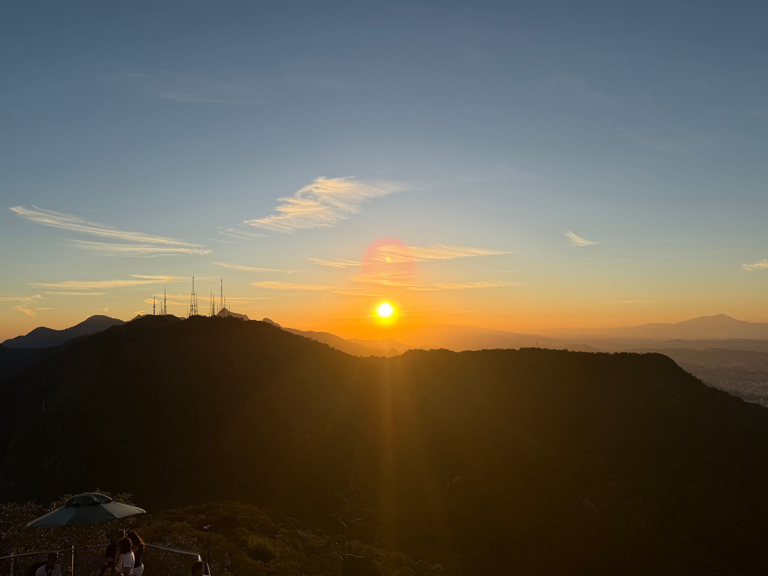 A sunset with a few wispy clouds in the sky, over a shadowy landscape of low rolling mountains, with several antenna towers on one peak.